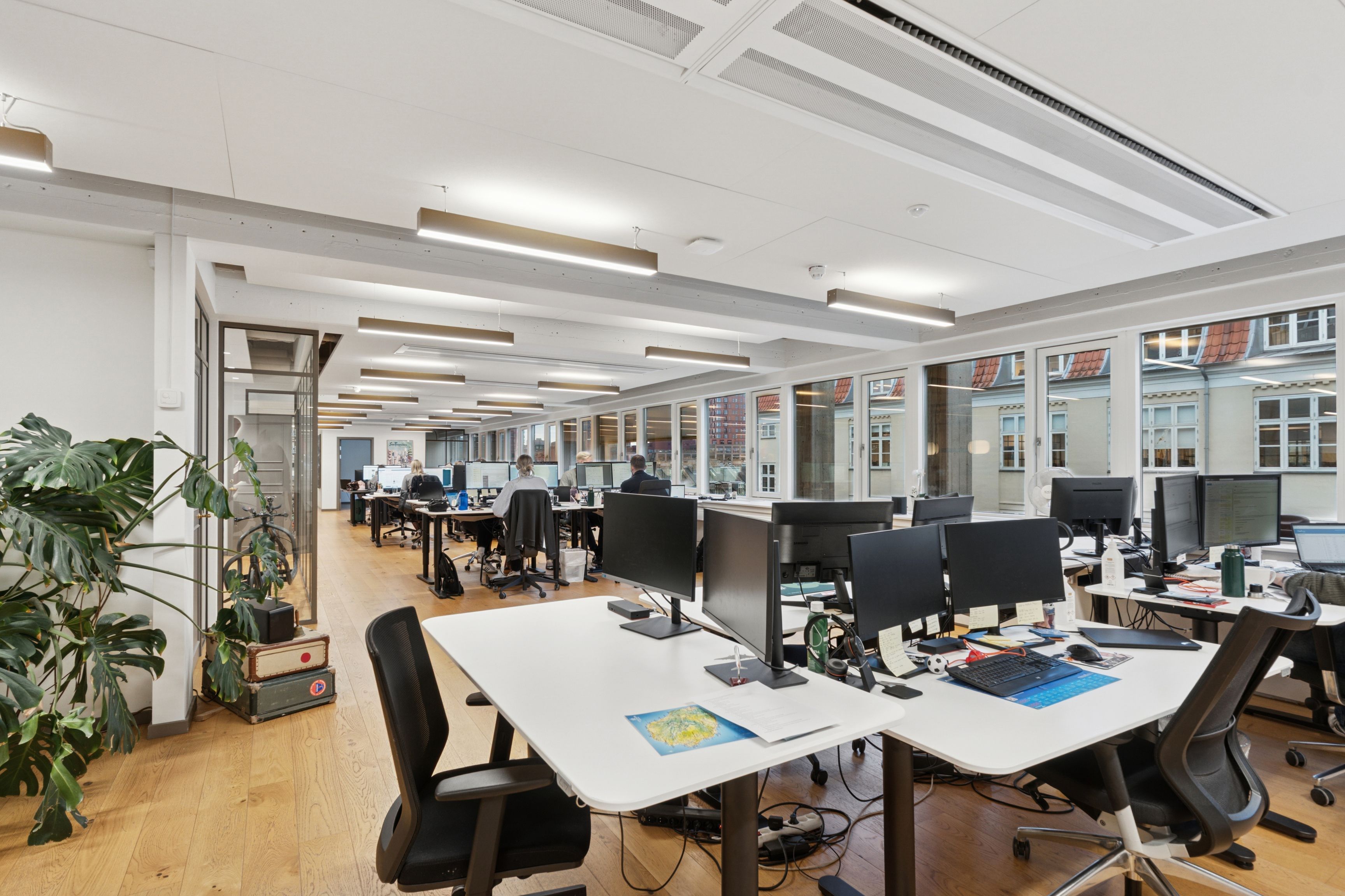 Kontorrum med højt til loftet og vinduer mod gaden og byen. Møbler, computere og planter er spredt rundt i lokalerne. Lysskiltene og ventilationskanaler er synlige i loftet.