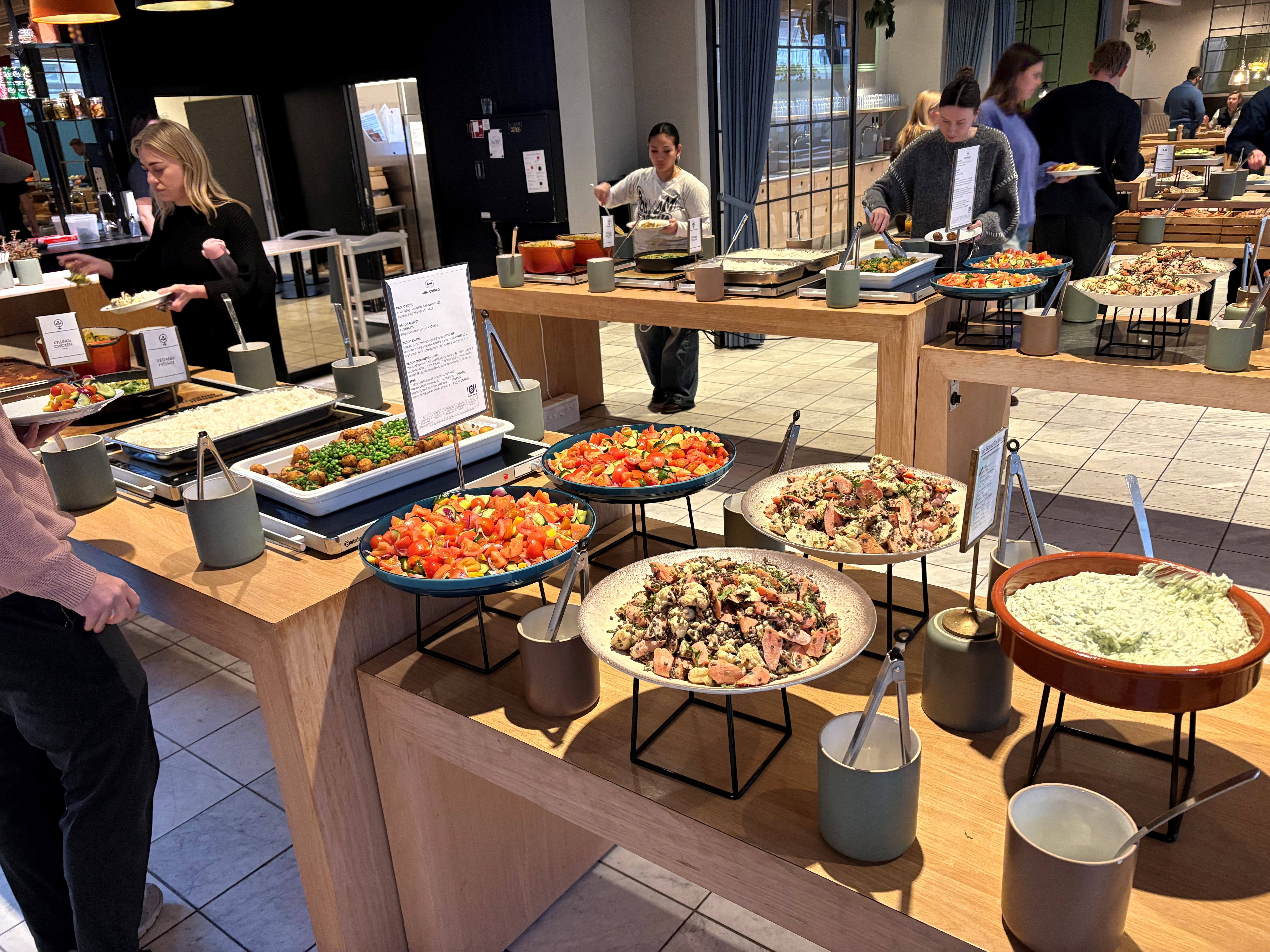 Diverse retter med salater og grøntsager i store skåle på flere serveringsborde i et moderne kontorområde med medarbejdere og glasvinduer i baggrunden