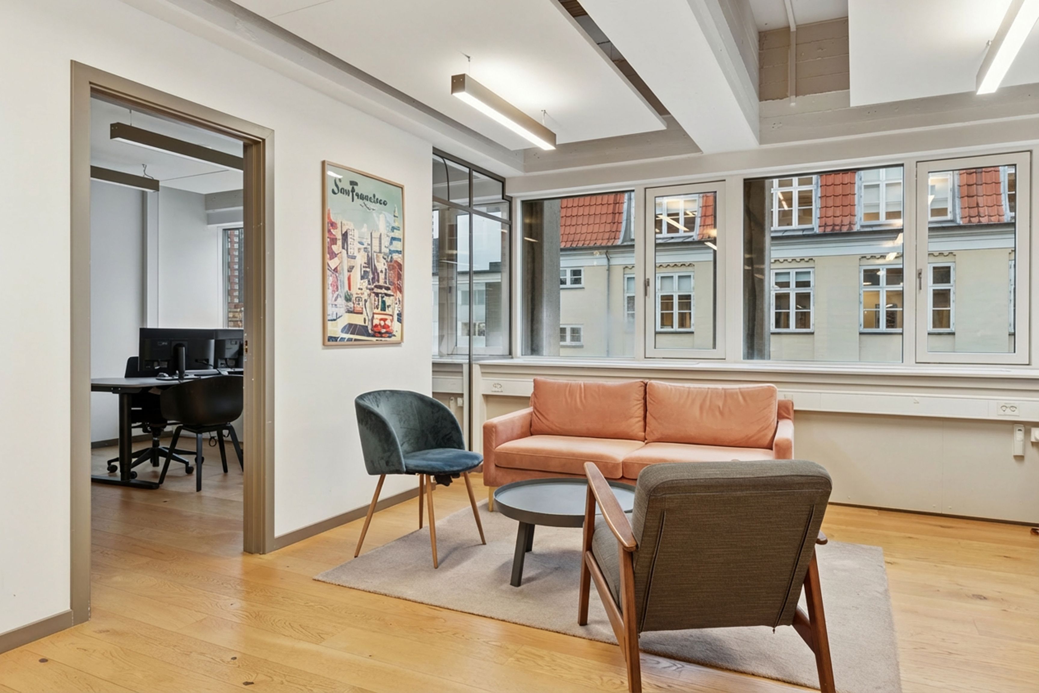 Kontorrum med brændenældeblå stol, lysebrun sofa, trægulv og store ruder med udsigt til en bygning i baggrunden