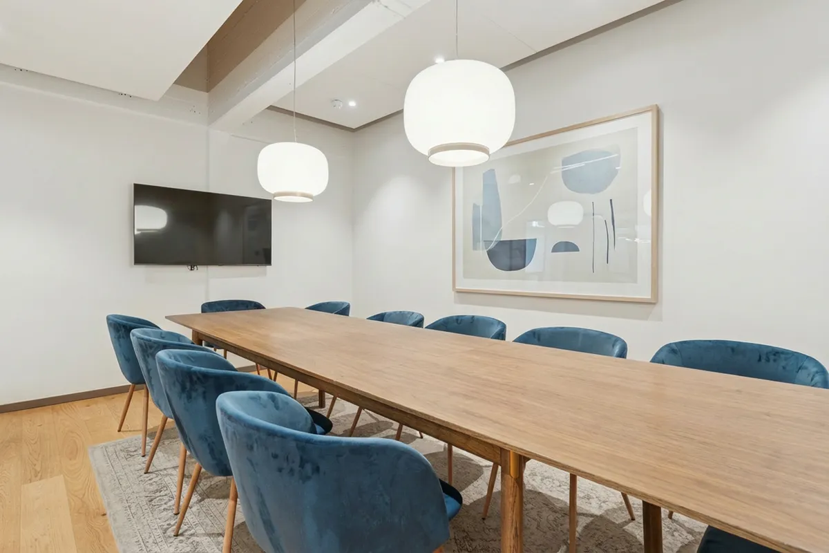 Kontorrum med langt bord og blå stolen i en stilfuld interiør, lyse kroner og tv-skærm på væggen, elegant gulvtæppe og abstrakt maleri