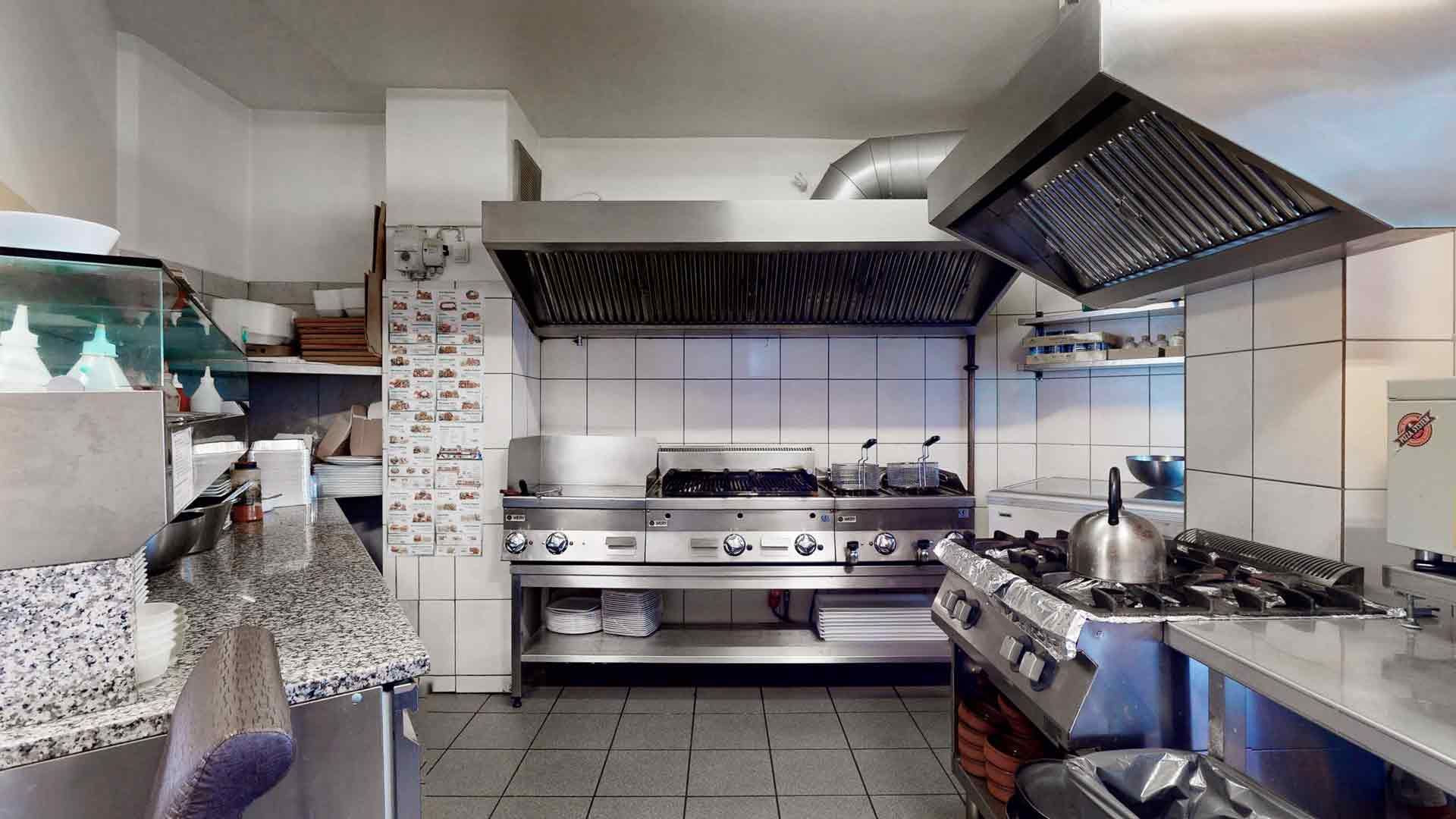 Et kommercielt køkken med moderne gaskomfur og elektrisk komfur samt ekstraktorer og stålskabe. Væggen er tømret med kakel og lysekroner er installeret på loftet.