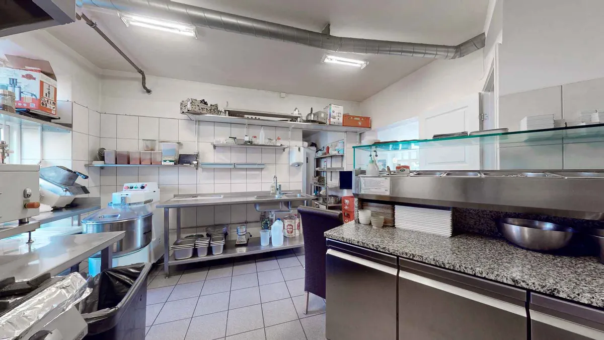 Et moderne og funktionelt køkken med lyse gulve, flere indbyggede apparater og en stor glasvitrine til udstødte retter