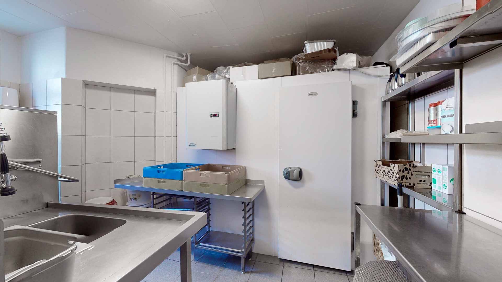 Et moderne køkken med flere stålborde, to vandhaner og en stålvaskedam. Væggene er hvide og der er et køleskab med et lager på toppen.