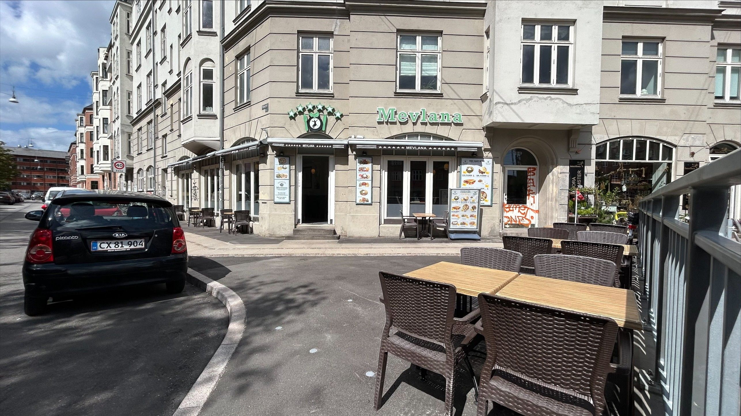 En restaurant med udendørs spisesteder og bord og stole på Jagtvej 119 i København. Bagved er en Kia Picanto parkeret og andre biler og bygninger