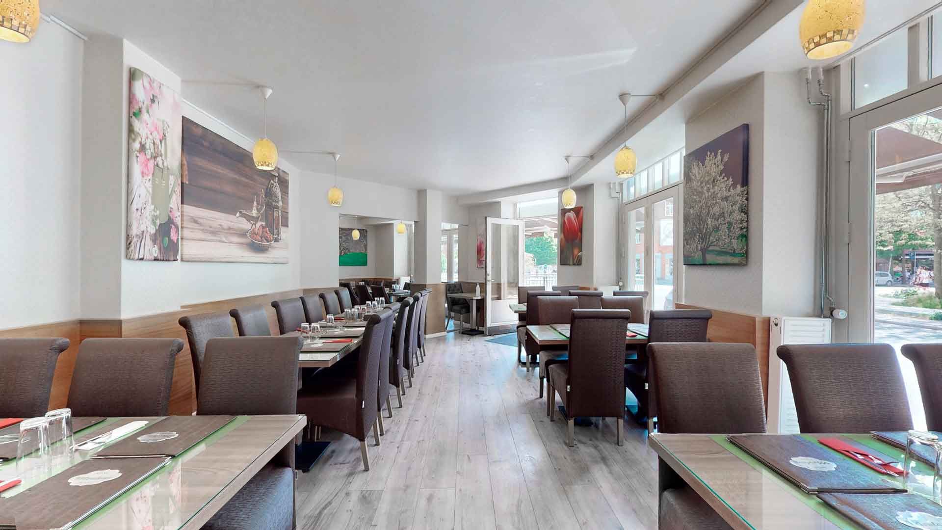 Restaurant med bord og stole, hvor bordene er dækket med menuer og glas. Væggene er dekoreret med malerier og lysekroner. Der er glasdøre og vinduer mod gaden.