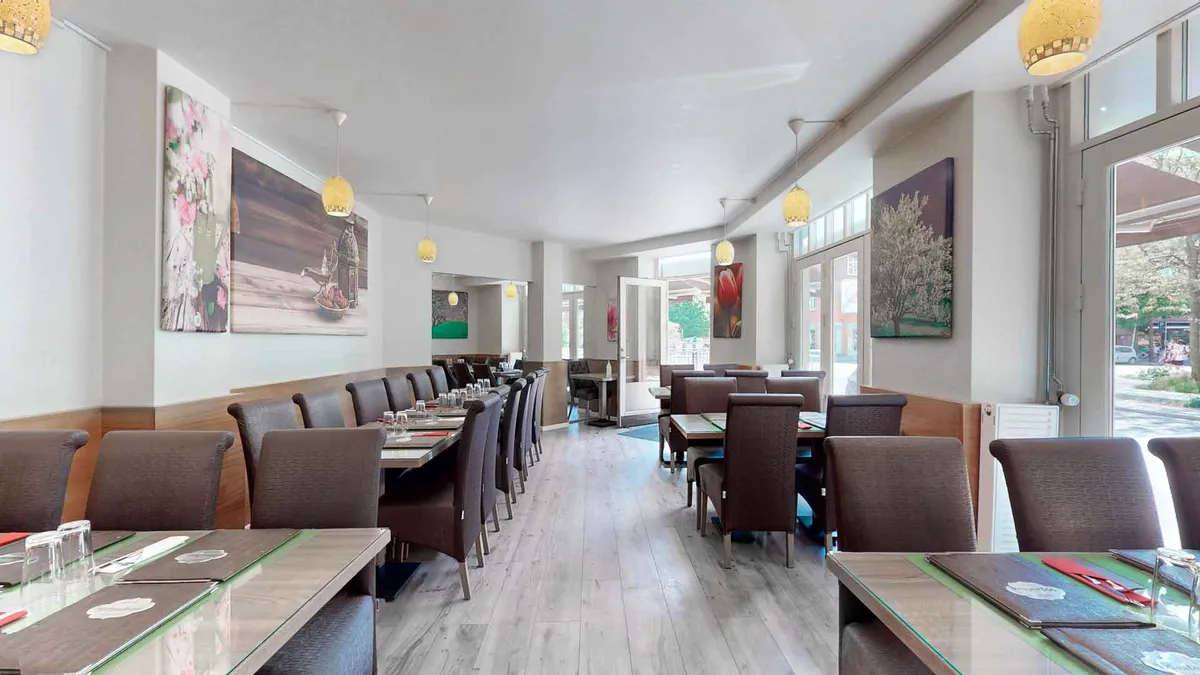 Restaurant med bord og stole, hvor bordene er dækket med menuer og glas. Væggene er dekoreret med malerier og lysekroner. Der er glasdøre og vinduer mod gaden.