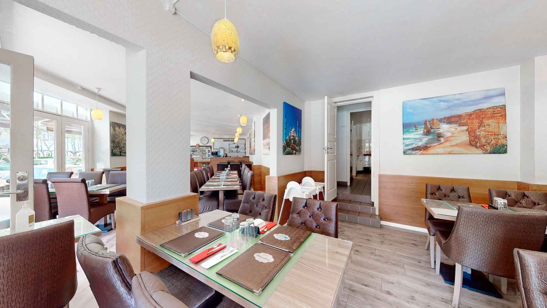 En restaurant med borde og stole, lysekroner, malerier og vinduer med udsigt til en have.