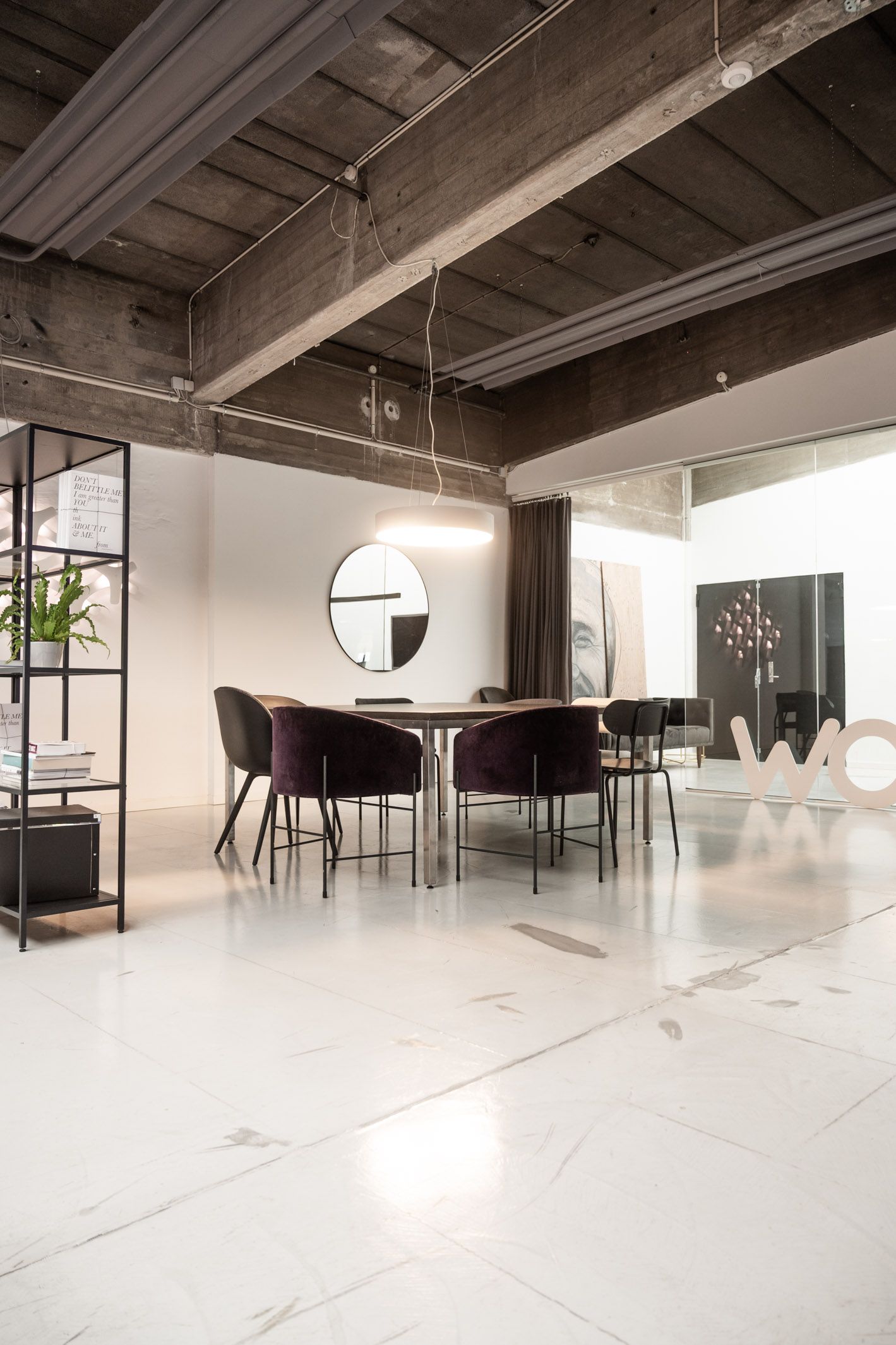 Rå og karakterfuldt kontor med marmorgulv og højloft. Rundt spejl og lysekroner hænger fra loftet. Bord med stole og plante samt møbler og logo i baggrunden.