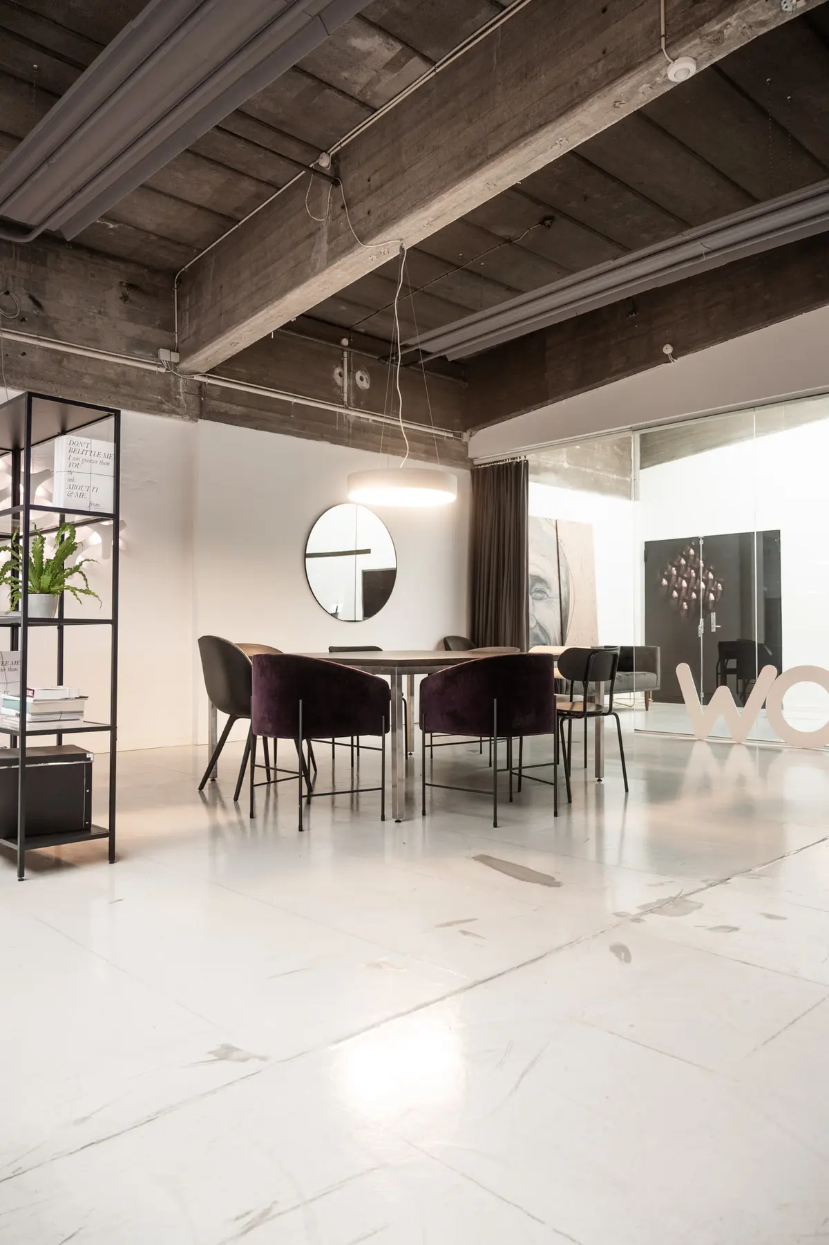 Rå og karakterfuldt kontor med marmorgulv og højloft. Rundt spejl og lysekroner hænger fra loftet. Bord med stole og plante samt møbler og logo i baggrunden.