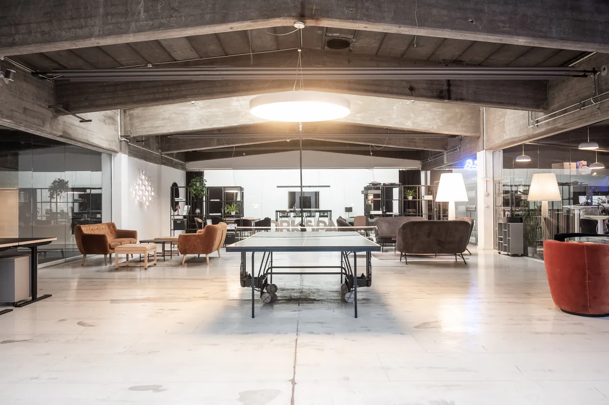 Kontor med pingpongbord og lyse lænestolpe i loftet med højde på ca. 4,5 meter. Rundt omkring står der bord og stole.