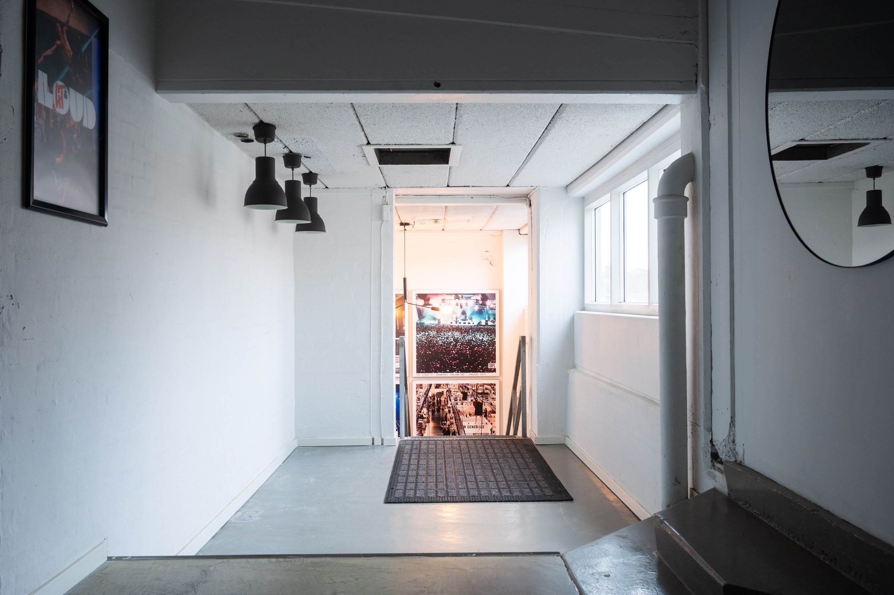 Københavns kontor med højt til loftet og lyse loftslampe i sort. Sort gulvtæppe foran døren og spejl med lyserefleks. Plakat med tekst og billede og en indgang til en anden lokal