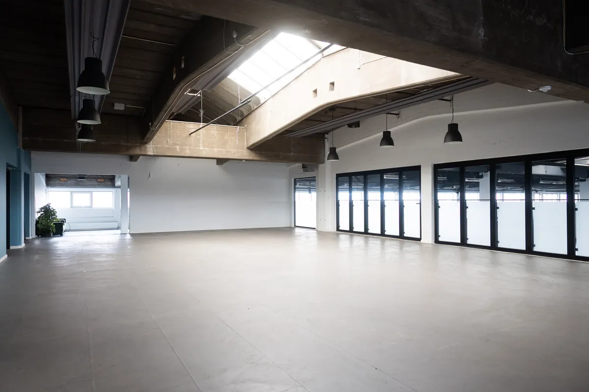 Kontor til leje på Raffinaderivej i København med højloft og store vinduer, lysekroner hængende og terrasse med plante. Betongulv og åben rum med udsigt til fabriksstrukturer.