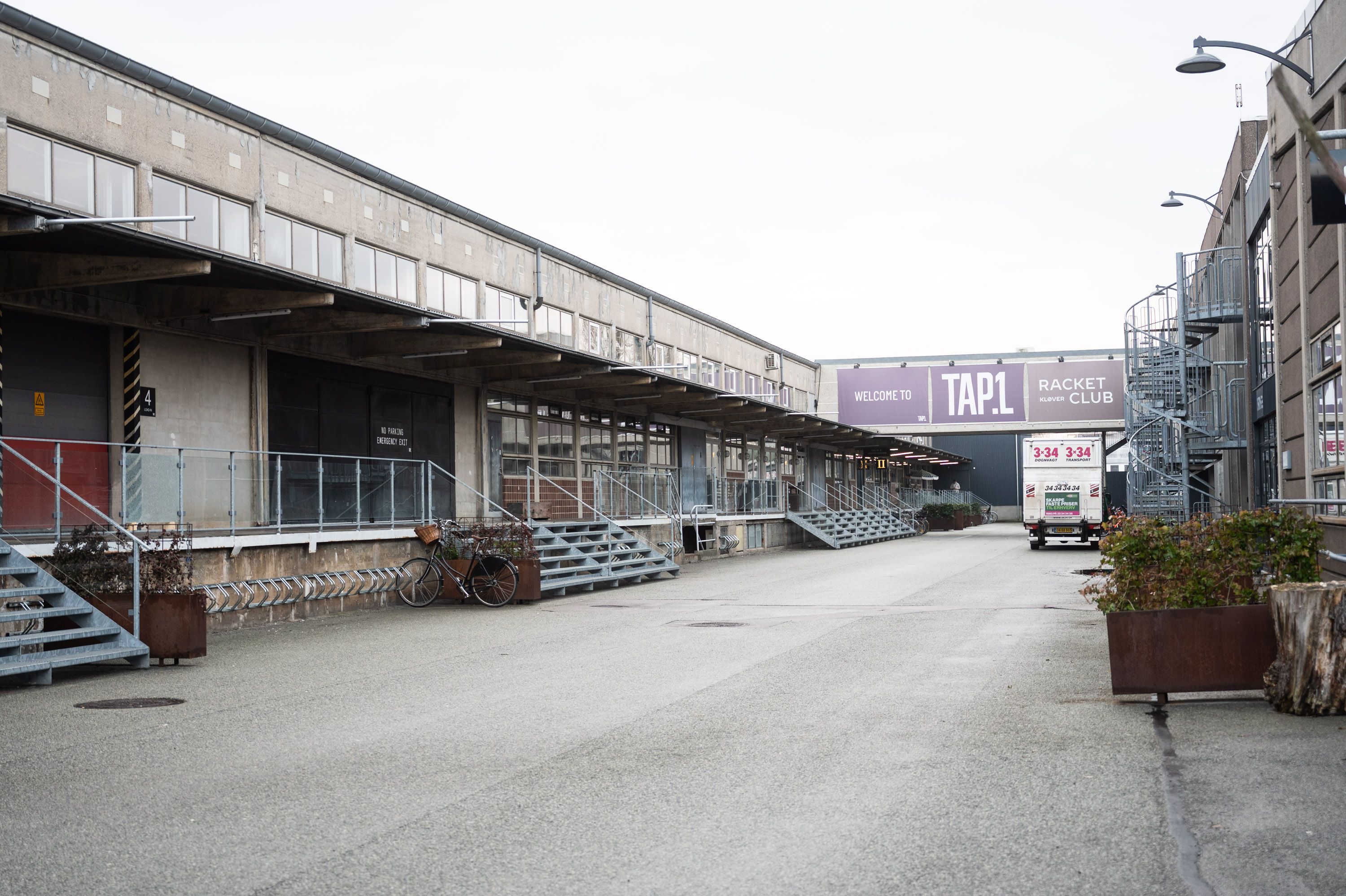 Råt kontor i TAP 1 med vinduer, gangbroer og en cykelparkeringsplads. Væggen med skiltet "Welcome to TAP 1" og "Racket Klub". En lastbil kører ud af parkeringspladsen.