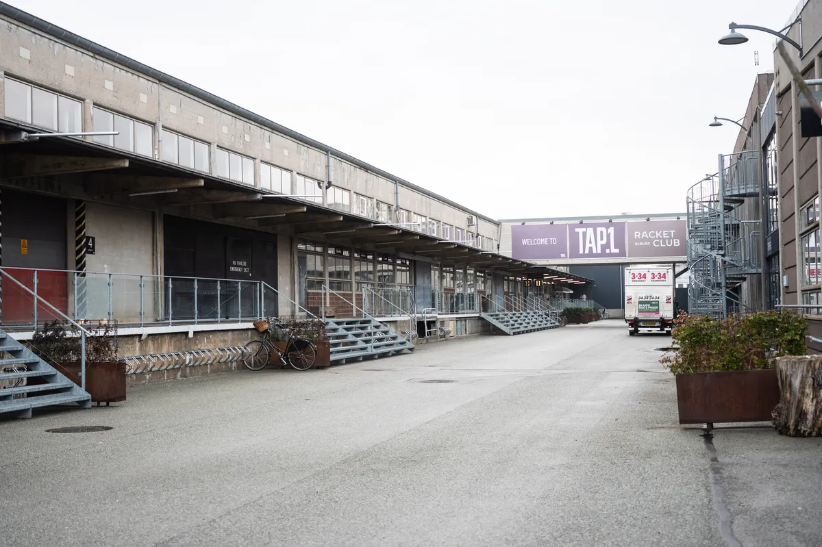 Råt kontor i TAP 1 med vinduer, gangbroer og en cykelparkeringsplads. Væggen med skiltet "Welcome to TAP 1" og "Racket Klub". En lastbil kører ud af parkeringspladsen.