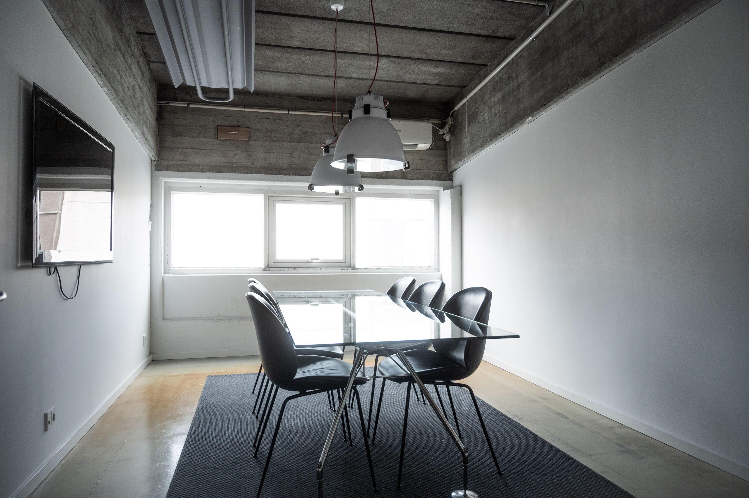 Kontor med glasbord og stolpelampe i loftet, hvor vinduer og hvid væg reflekterer lys. Tæppe og møbler er placeret under vinduet.