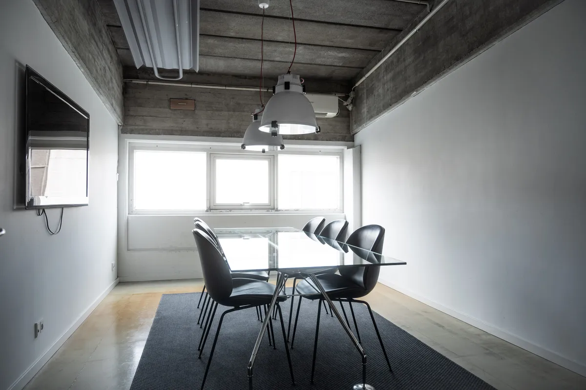 Kontor med glasbord og stolpelampe i loftet, hvor vinduer og hvid væg reflekterer lys. Tæppe og møbler er placeret under vinduet.