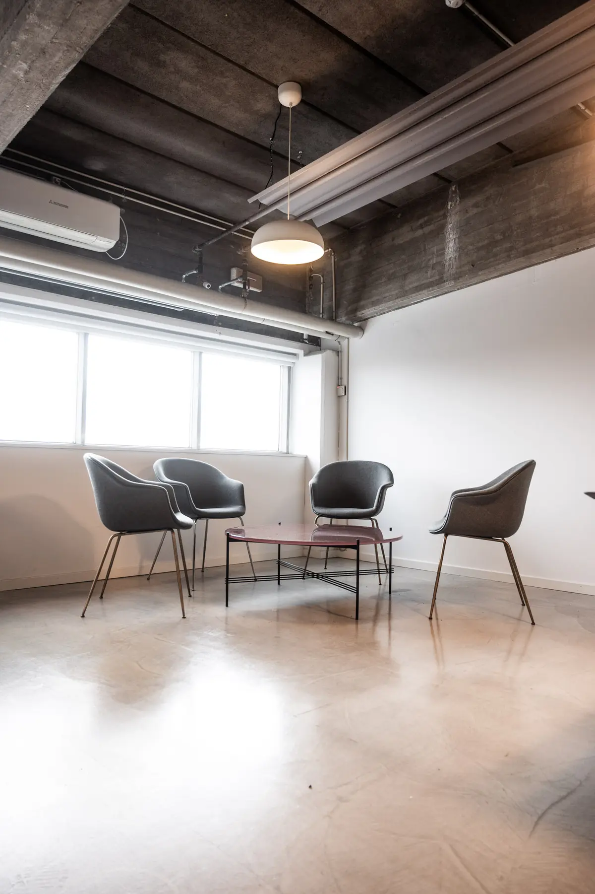 Kontorrum med højloft, fire stole, rødt træbord og lysekroner. Hvide vægge og vinduer samt klimaanlæg.