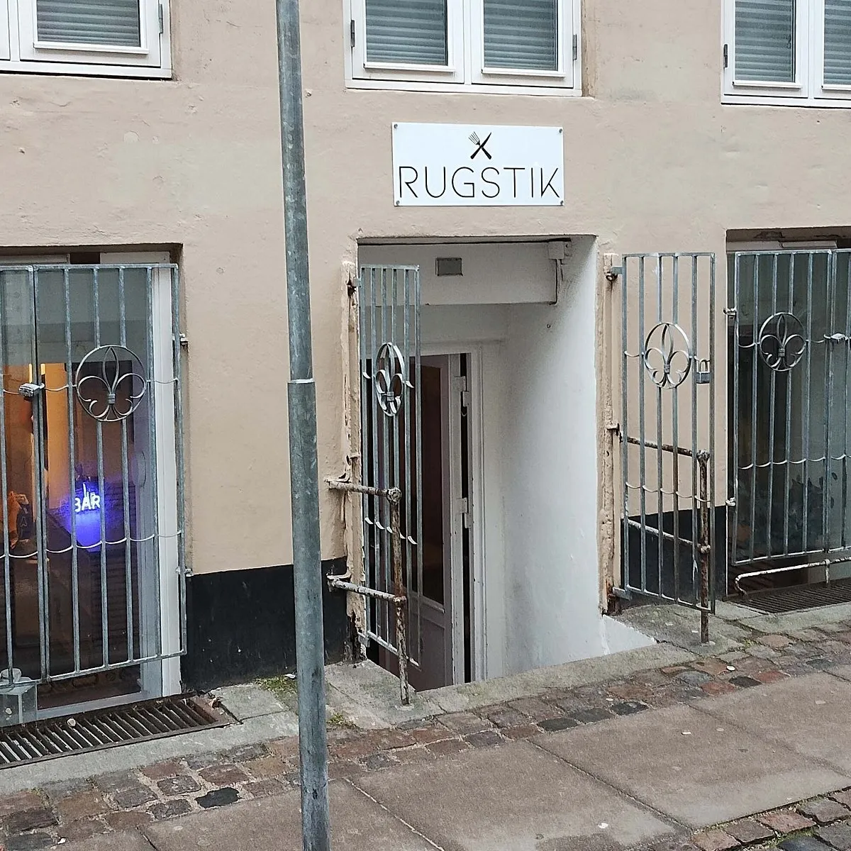Rustne metaldøre med låse og udsigt til café med 3 Bar-skilt, placeret på en stenbrolægget gågade i København