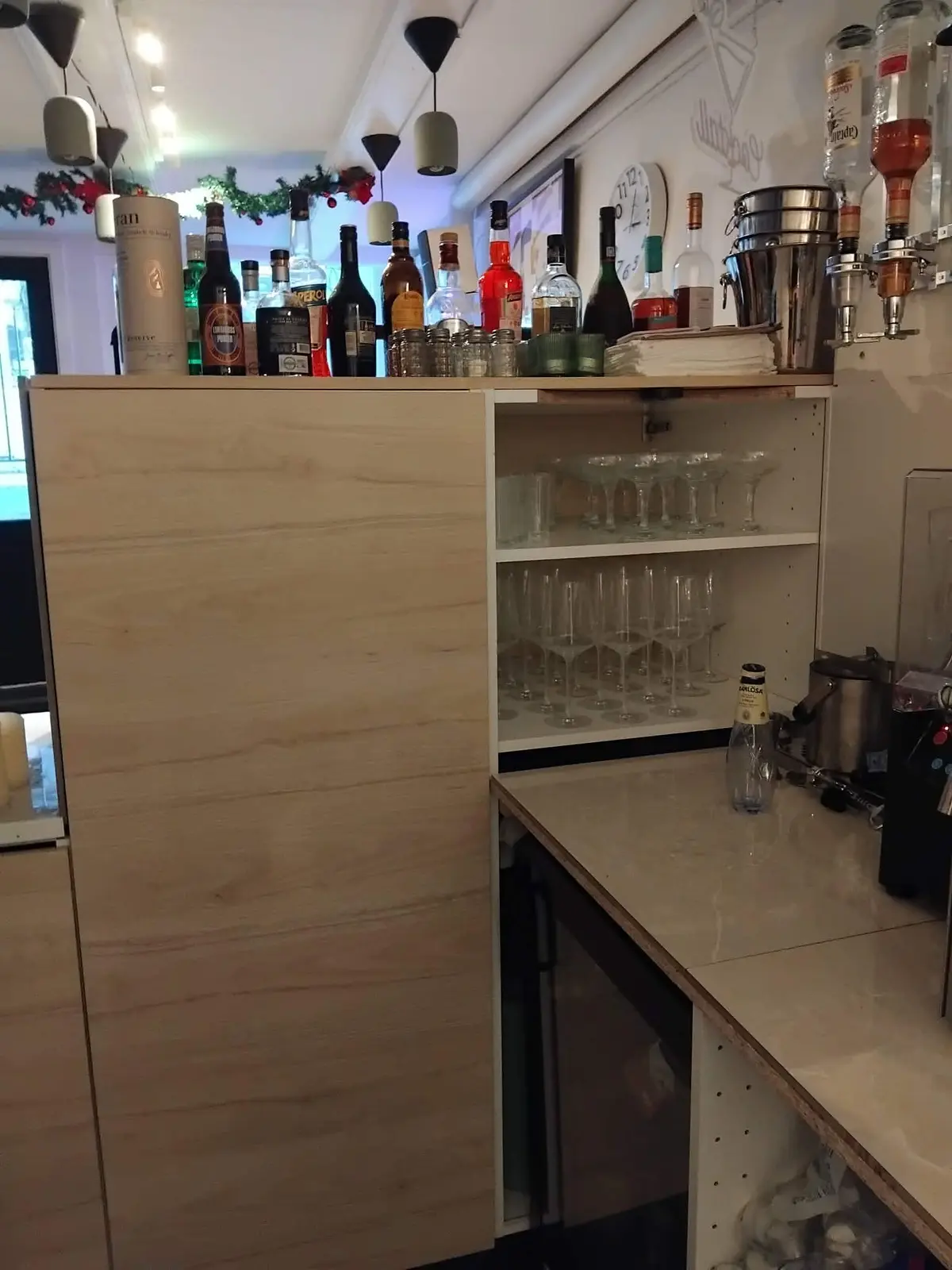 Hyggelig café med åbent køkken og bar med øl, vin og spritflasker samt glas og drikkeglas samt champagne i køleskab og flasker på hylde