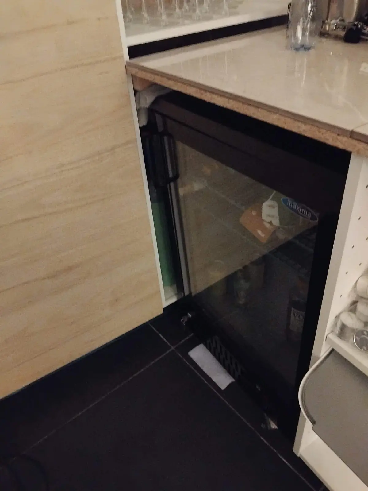 Under en køkkenbord er der et køleskab med etiketter og drikkevarer. Der er også en stang med kopper og en åben skuffe med en håndklæde.