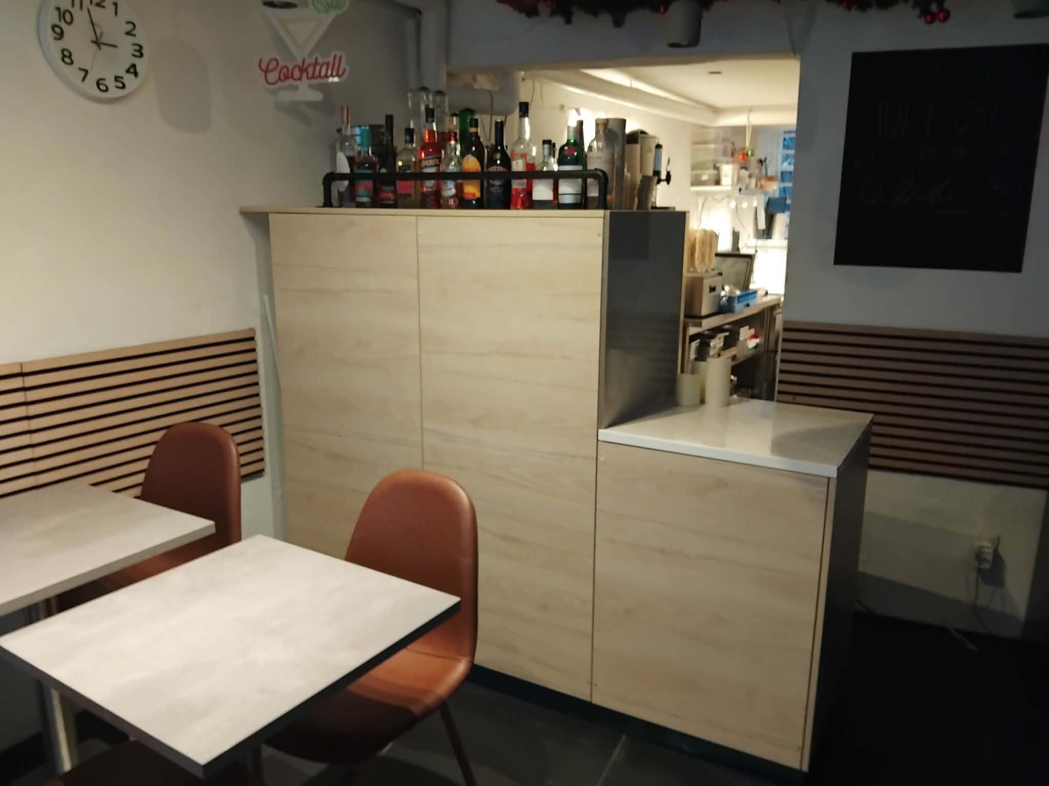 I caféen er der træborde, brune stole og en lyse træskab med hylder, hvor der er placeret glasflasker. På væggen er der et ur og en tekst, der læser "Cocktail"