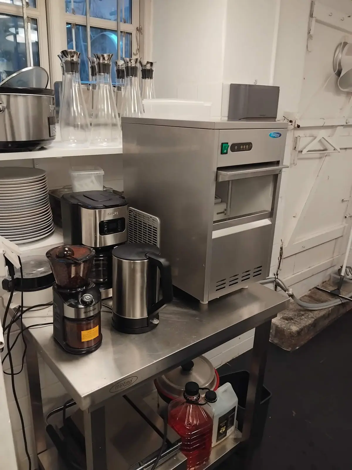 Køkken med ismaskine, kaffemaskine, kaffetrakter og flasker på en stor serviervogn med flere apparater og tallerkener