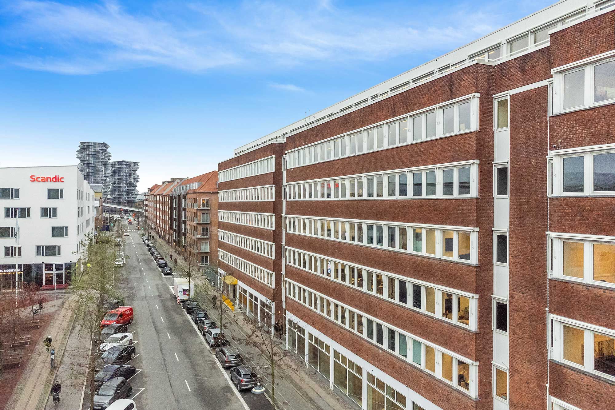 Kontorlejemål med panoramaudsigt over Kødbyen og privat terrasse. Bygningen med mange vinduer og moderne design. Parkerede biler langs gaden og bygninger i baggrunden.