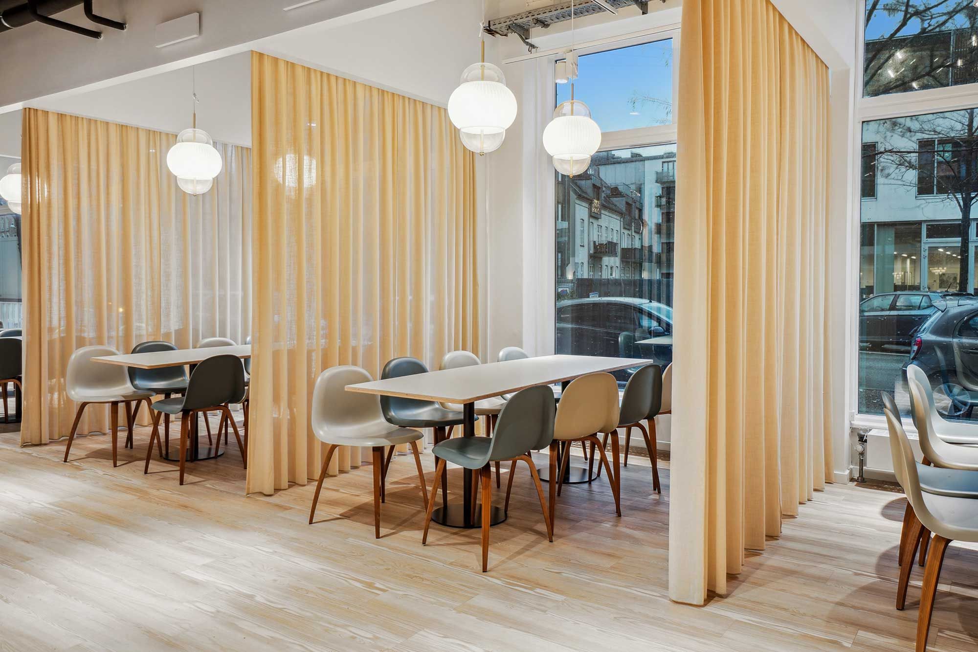 Kontorrum med lyse gulve og hvide vægge, runde borde og stole, lysekroner, og guldbrune gardiner, der stiger op til loftet. Der er udsigt til byen gennem store ruder, hvor biler og bygninger er synlige.