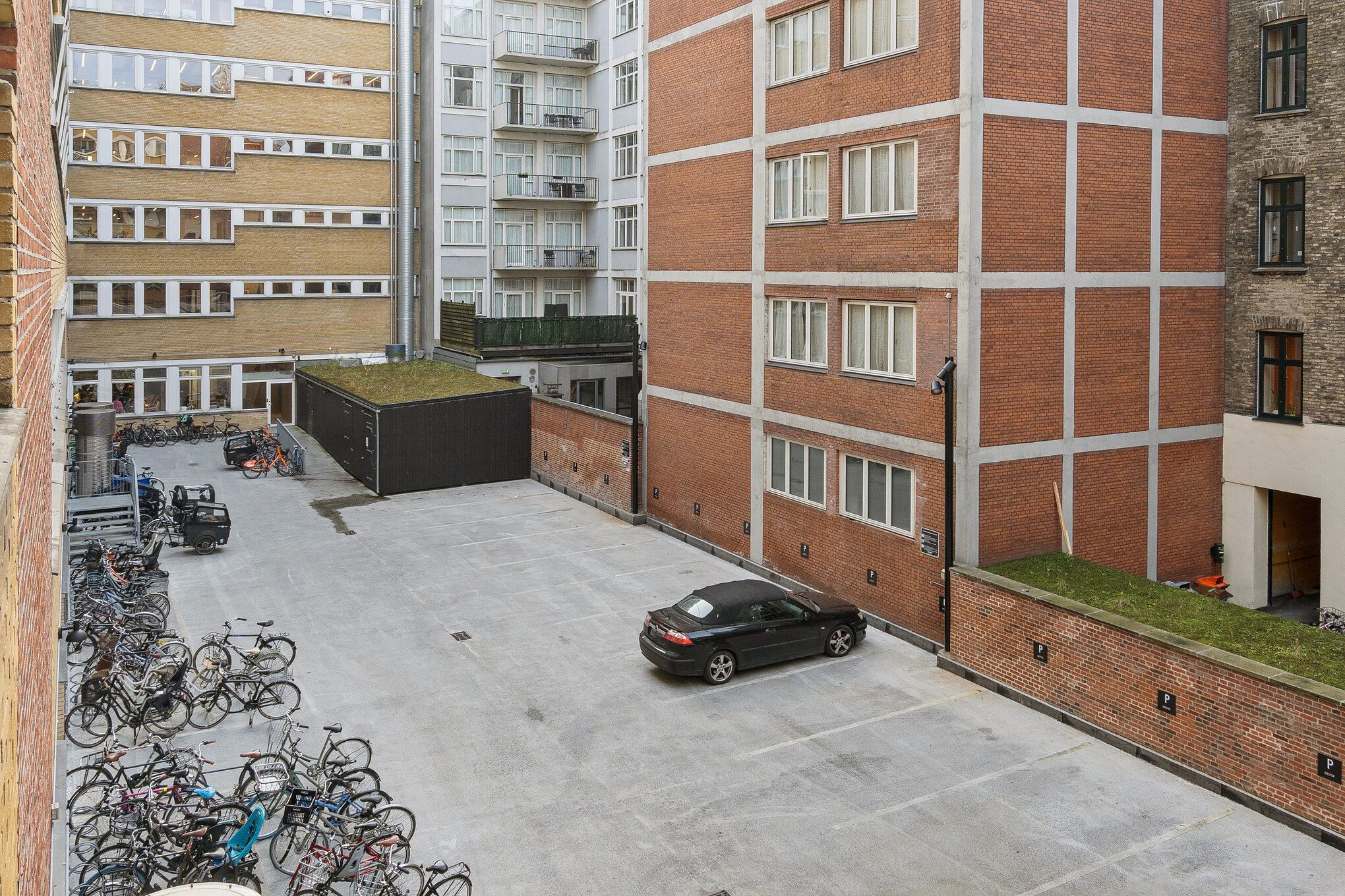 Parkeringsplads med parkerede biler og cykler i en bygning med mange vinduer og balkoner, samt en grøn taghave