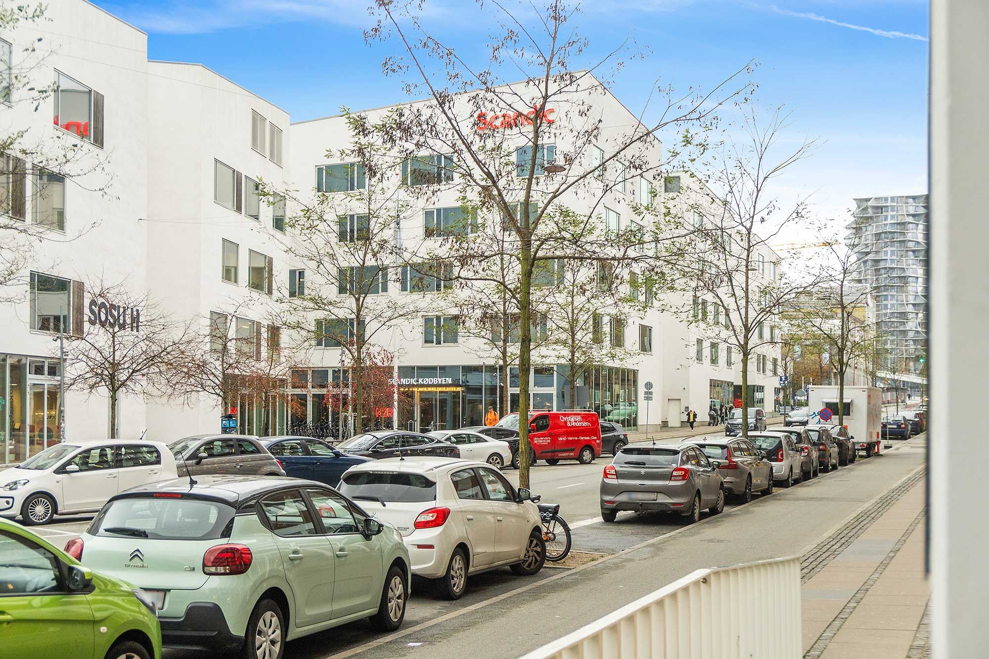 Kontorhus med mange vinduer og skandic-logo. Parkerede biler og folk går på fortovet. Bygning med terrasse og udsigt over Kødbyen i København.