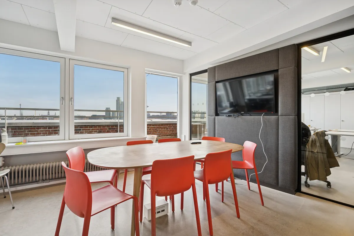 Kontorlokaler med moderne møbler og en stor panoramaudsigt over byen. Rundt bord med røde stole og en tv-skærm. Et lysere gulv og hvide vægge.