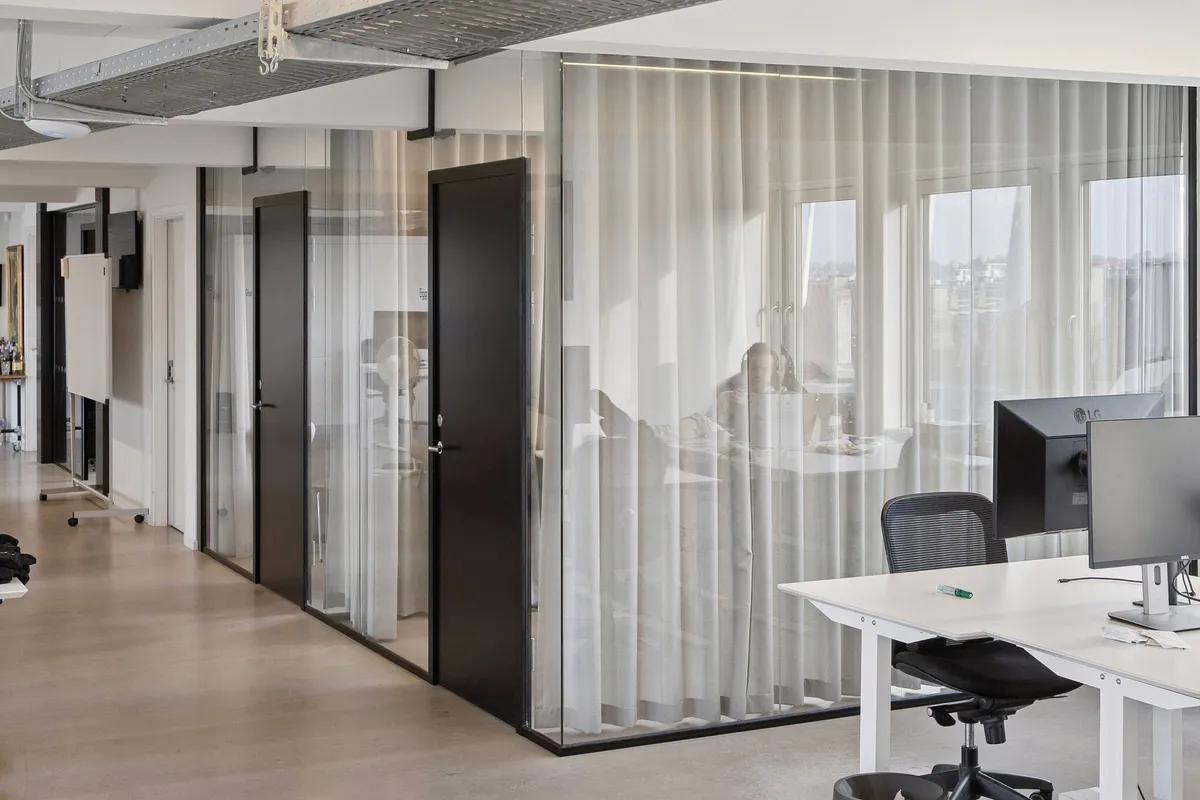 Kontor med store vinduer og terrasse med udsigt over byen. To monitorer på et bord med en stol og en skraldespand i nærheden.