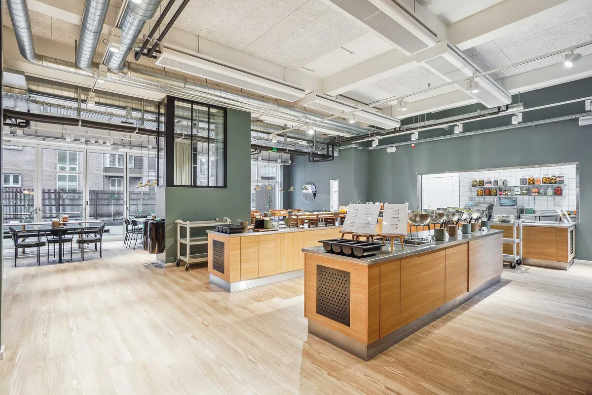 Kontor med højt til loftet og lyse loftsvinduer, hvid loftsbjælke og møbler med grønne vægge, trægulve og bord og stole samt terrasse med udsigt over byen