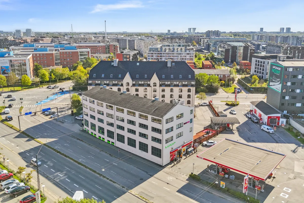 Kontorbyggeri med flere etager og vinduer med udsigt over København, to biler parkeret, et stort antal biler og folk på vejen, et pumpestation og et reklameskilt