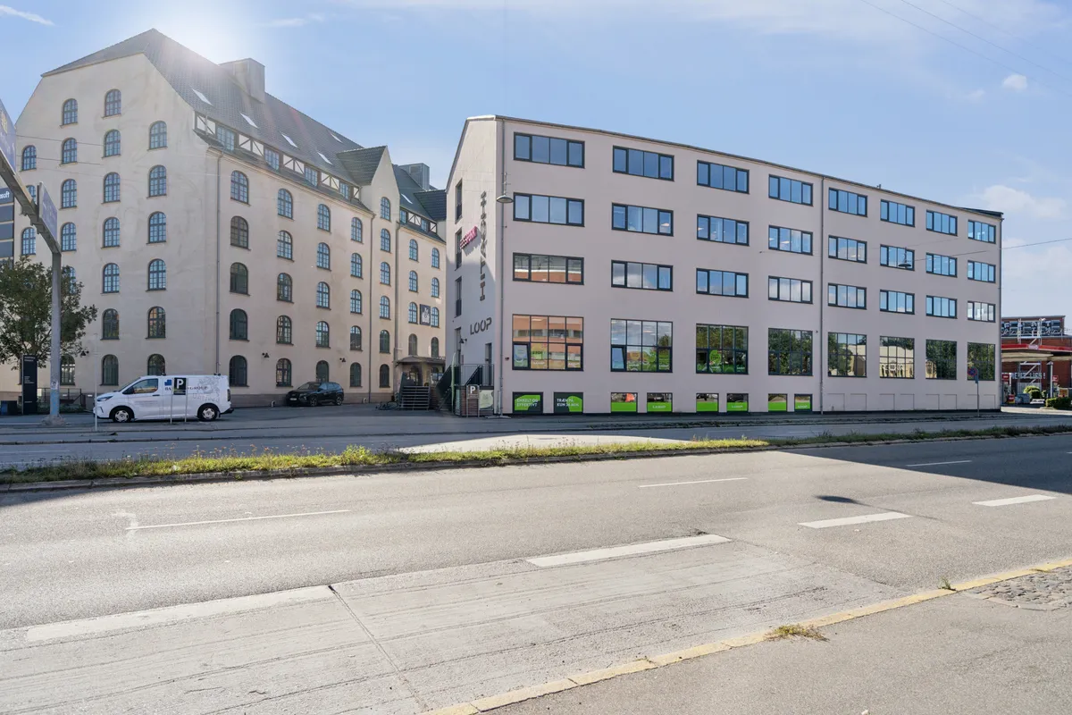 Kontorbyggeri med lyse vinduer og lysegrå facader, hvor en hvid lastbil parkeret på gågaden foran, og et rødt bygning i baggrunden