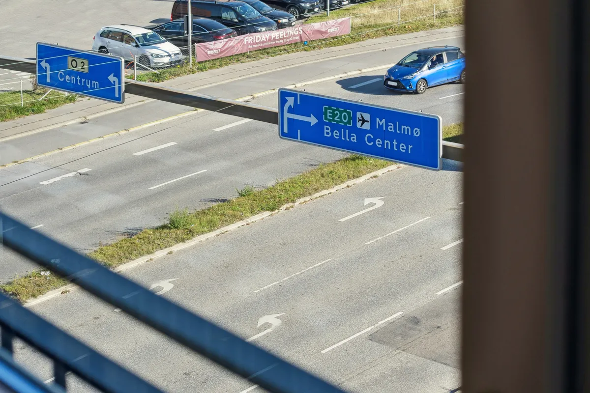 Se et parkeringsområde med biler parkeret, hvor en bil kører, og der er skilte til Centrum og Malmø Bella Center