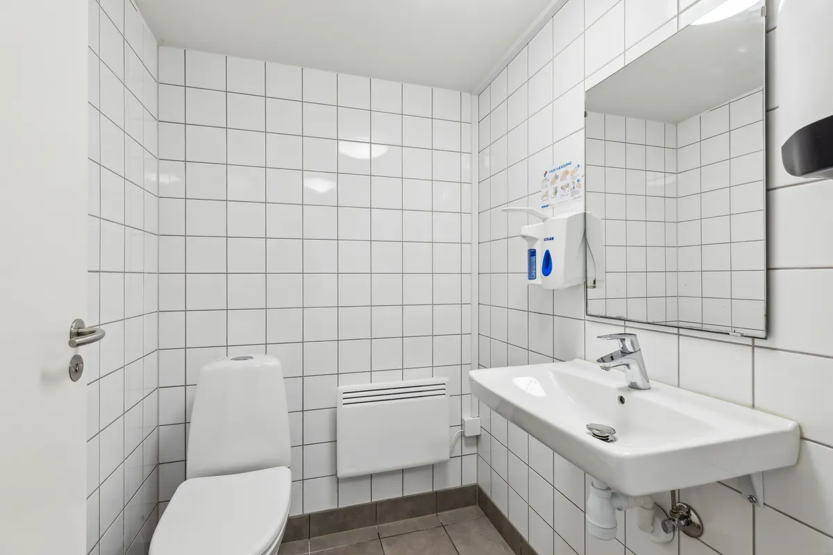 En hvid toilet, hvidt badeværelse med hvidt spejl og hvidt toiletpapir dispenser på væggen, en hvid vandhaner og et hvidt vask