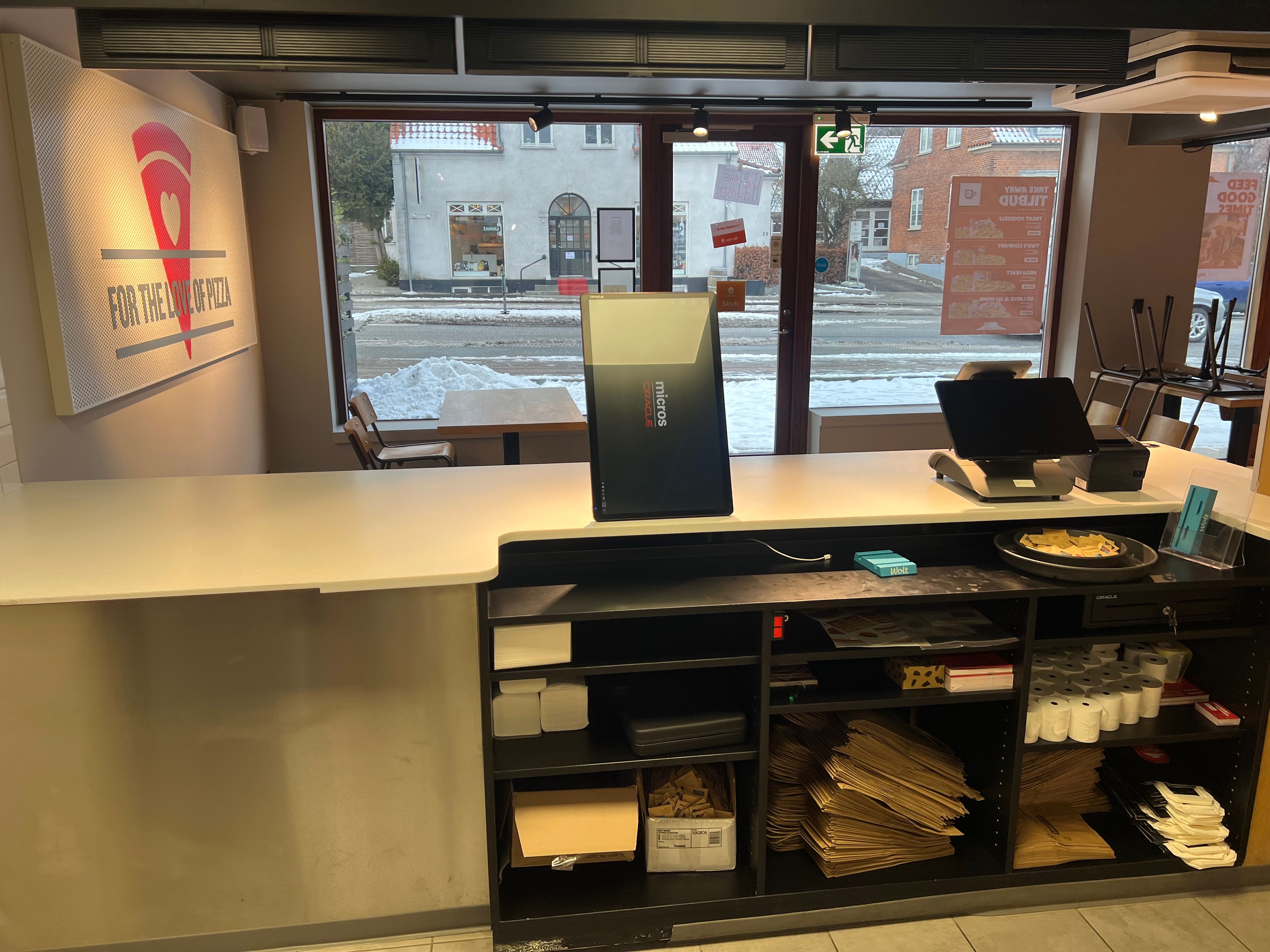 Take-away med serveringsbord, kasseapparat og salgsskilt med forretningsnavnet. Papirposer og kasser er placeret under bordet.