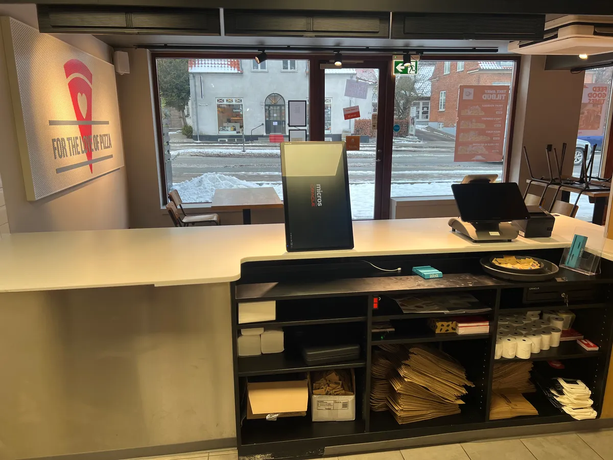 Take-away med serveringsbord, kasseapparat og salgsskilt med forretningsnavnet. Papirposer og kasser er placeret under bordet.