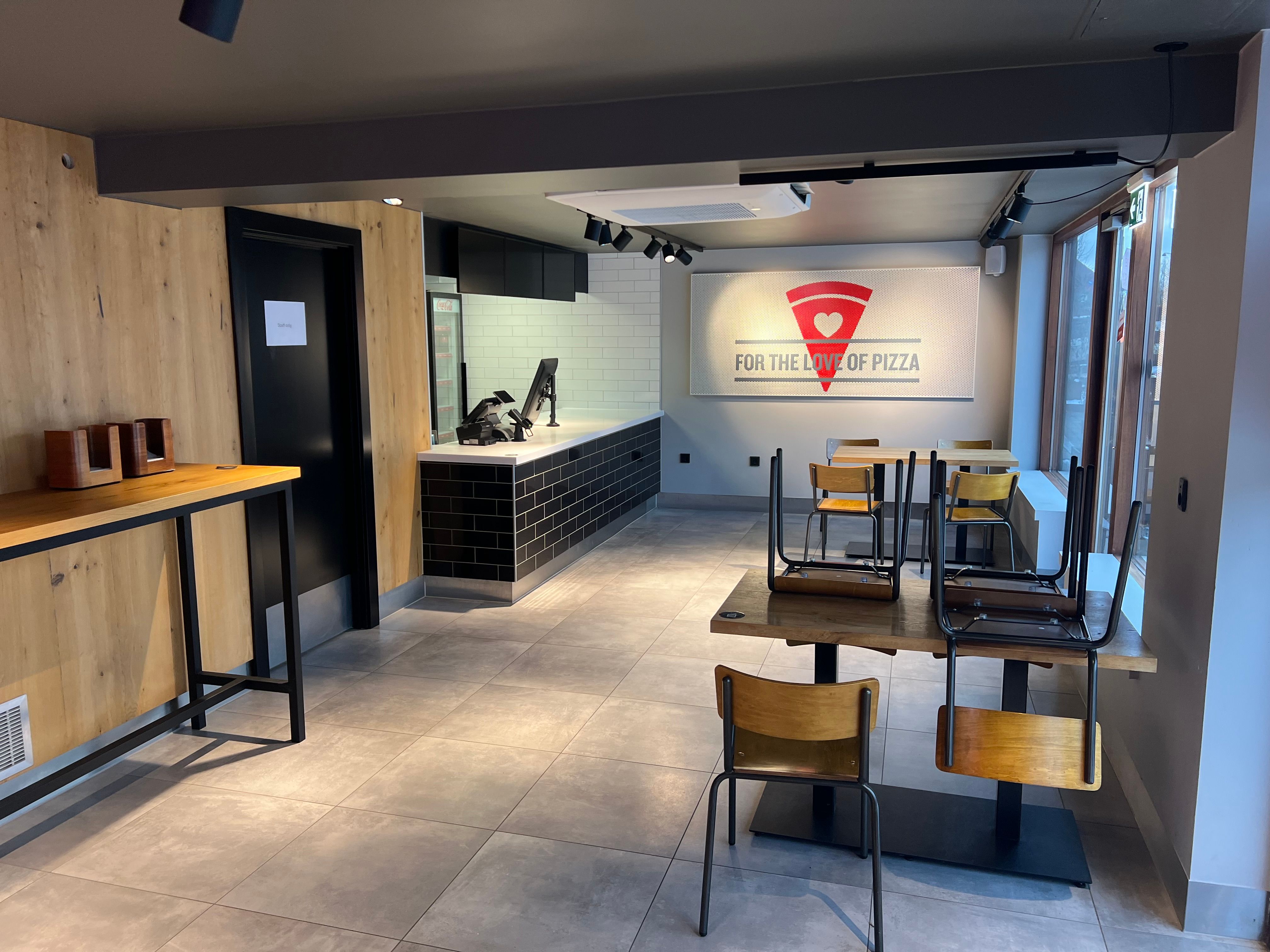 Take-away med store vinduer og træpaneler. To lange borde med stole og skrift, der siger For the love of pizza.