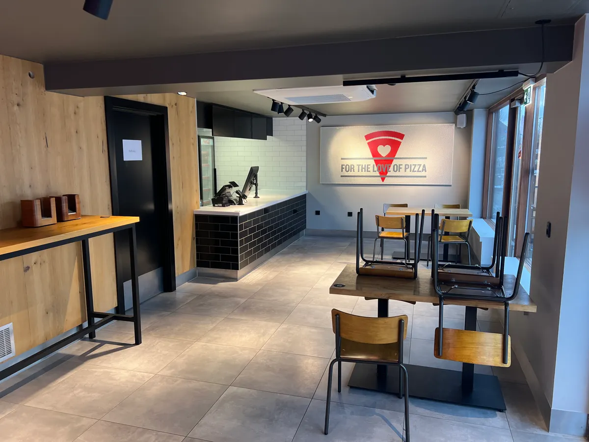 Take-away med store vinduer og træpaneler. To lange borde med stole og skrift, der siger For the love of pizza.