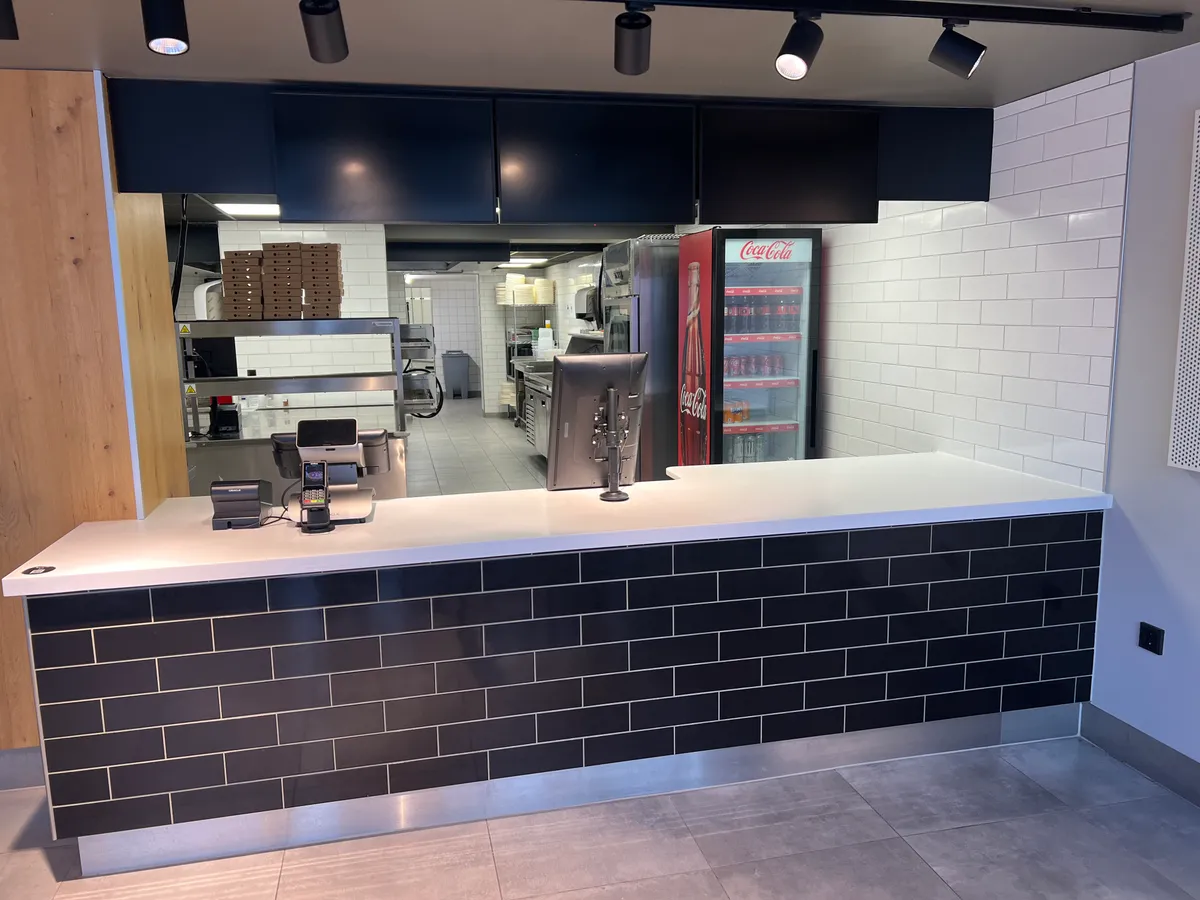 Take-away med serveringsbord og bestillingsterminal. I baggrunden er der et køleskab med Coca Cola og et køkken med flere apparater.