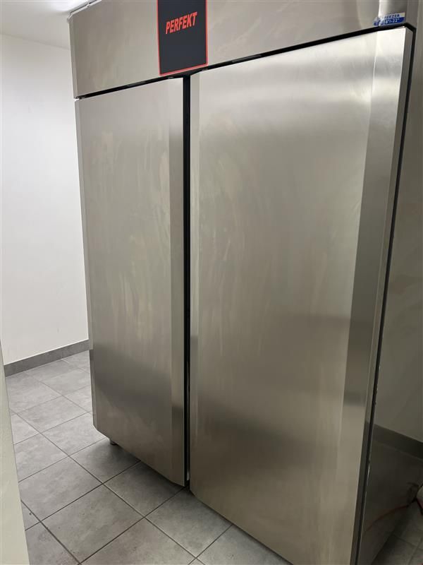 En stor, sølvfarvet køle- og fryseskab med tekst og logo i højre side. Skabsdørene er åbne, og gulvet er lavet af lysegrå fliser.