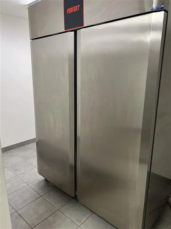 En stor, sølvfarvet køle- og fryseskab med tekst og logo i højre side. Skabsdørene er åbne, og gulvet er lavet af lysegrå fliser.