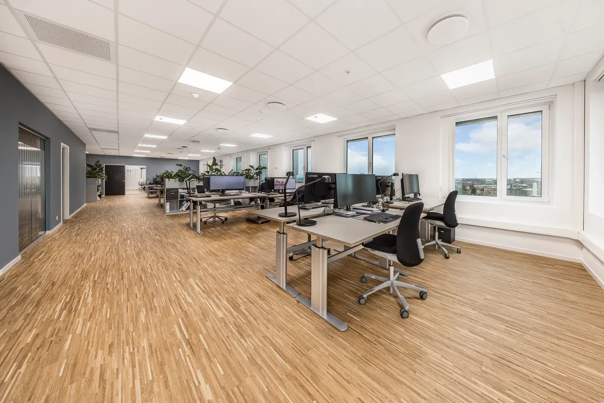Kontorområde med højloftede rum og lyse gulve. Værelser med borde, stole og computere samt plantager og vinduer med udsigt til byen.
