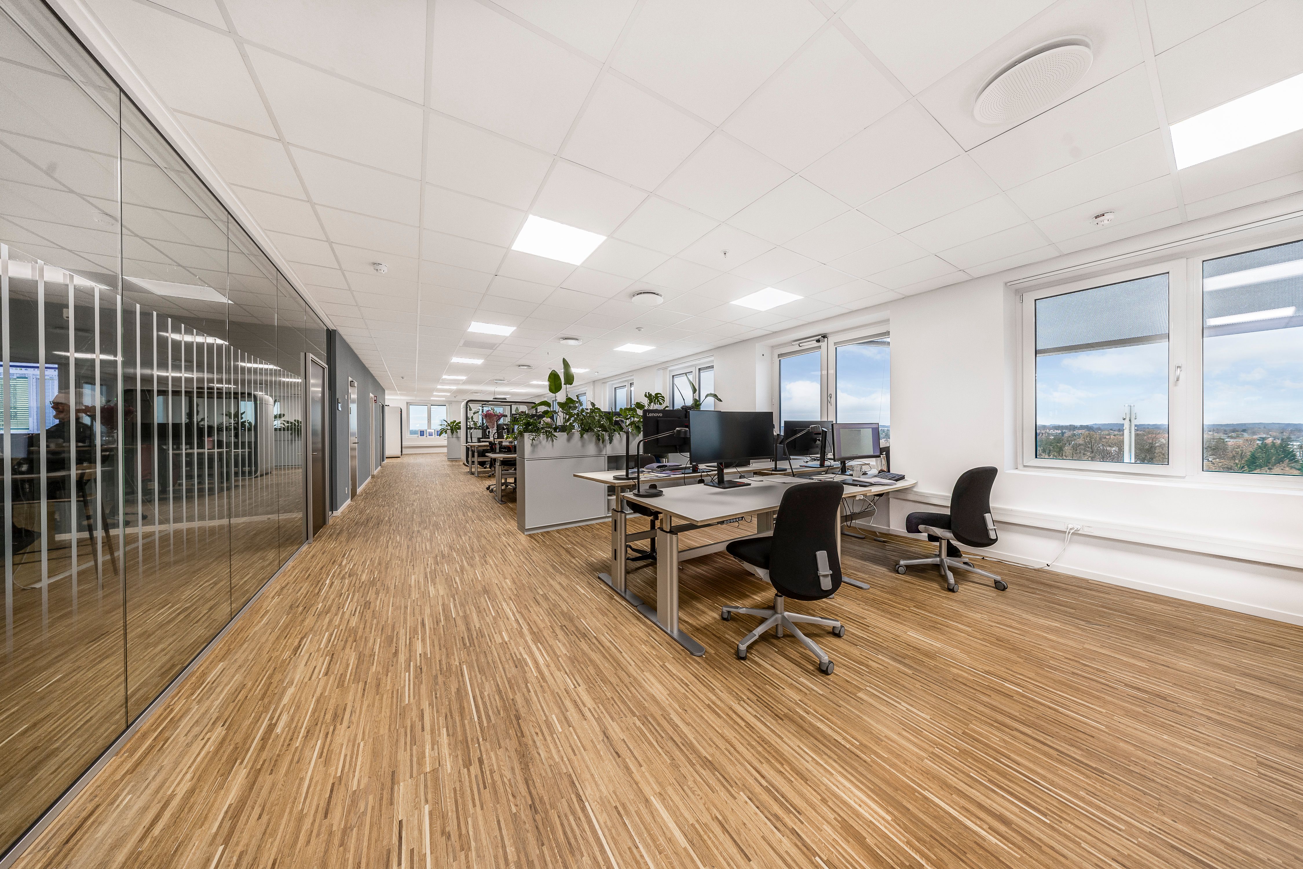 Elegant kontor med lyse gulve og store ruder med udsigt over byen. Kontorbord med computere, skrivebordslamper og to sæder. En person står ved et andet skrivebord.