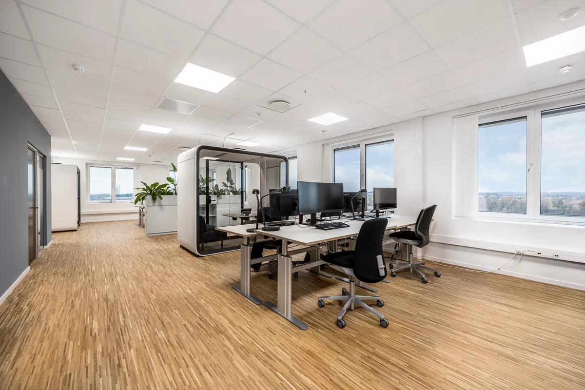 Kontor med høj service og smukke fællesområder. Store vinduer, moderne skriveborde og plante. Hvid gulv og vægge. Et lukket kabinet med plante og skrivebord.