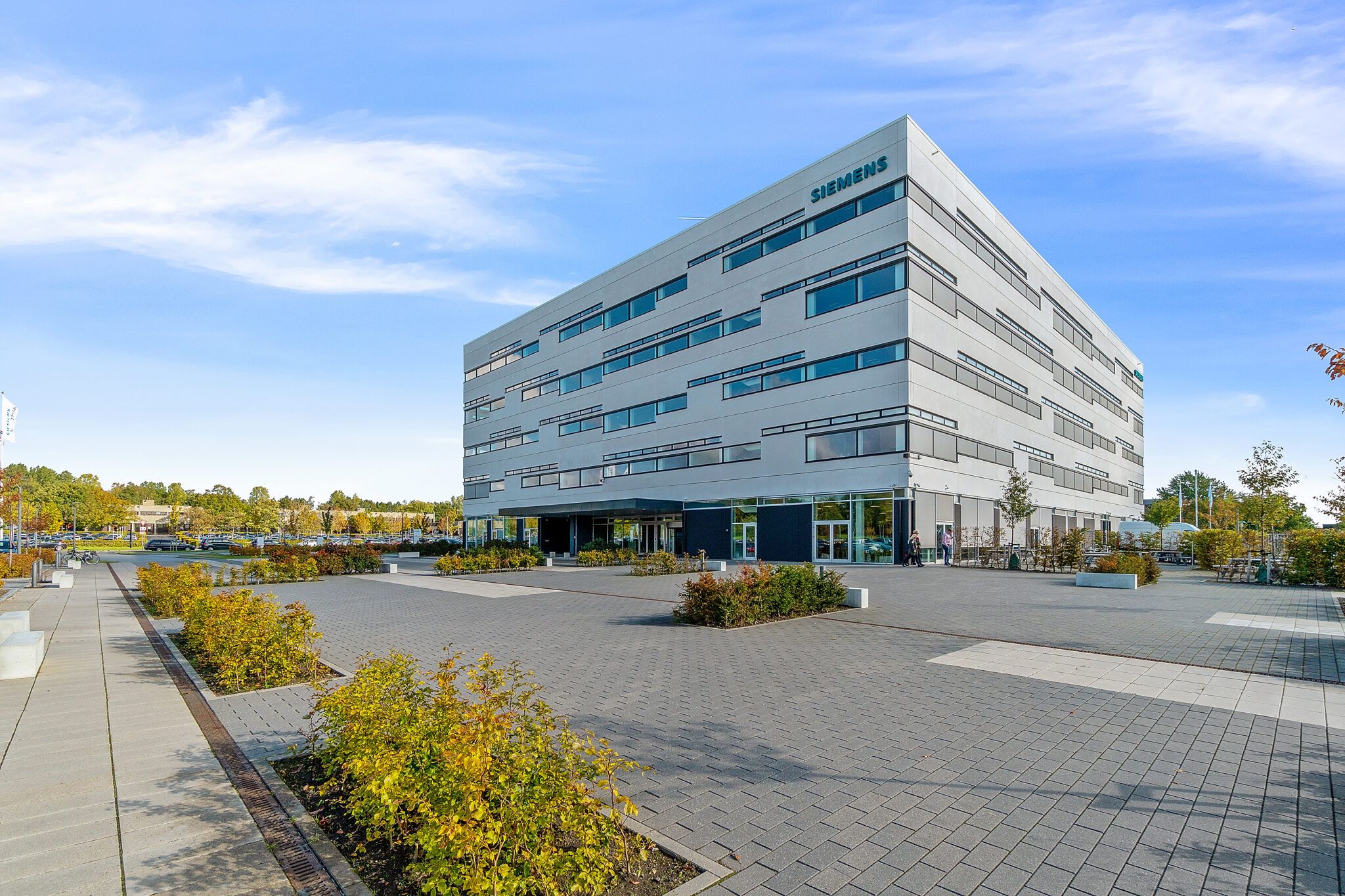 Siemens kontorlokaler med stort parkeringsområde og grønne anlæg. I baggrunden er der skov, og det er en solrig dag med skyer på himlen
