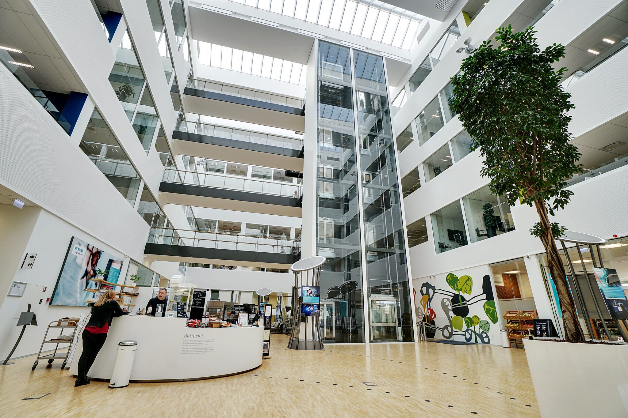 En moderne lobby med høje glasvægge, en stor hvid plante og et køkken med en mand bag disken og en kvinde foran