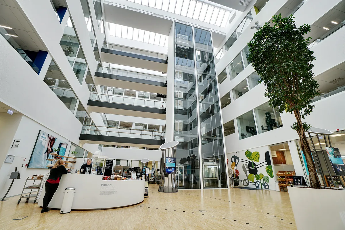 En moderne lobby med høje glasvægge, en stor hvid plante og et køkken med en mand bag disken og en kvinde foran